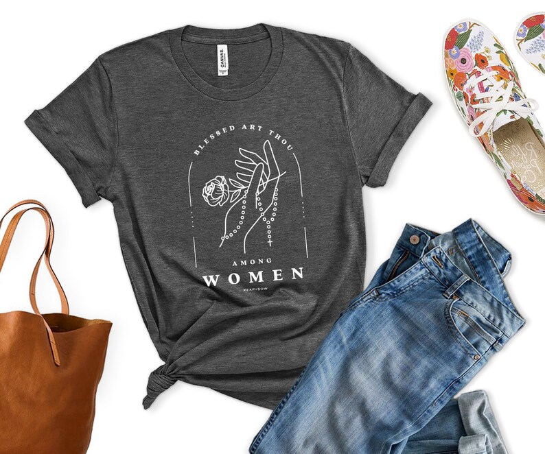 Peut inclure: Un t-shirt gris fonc&eacute; avec un motif blanc repr&eacute;sentant des mains tenant un chapelet et une fleur. Le texte "Blessed Art Thou Among Women" est imprim&eacute; sous le motif.
