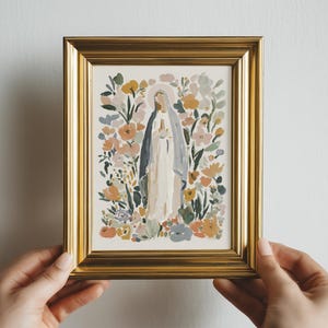 May include: A framed art print featuring a religious figure surrounded by colorful flowers. The artwork is in a gold frame and held by hands. The figure is wearing a white robe and a blue veil.