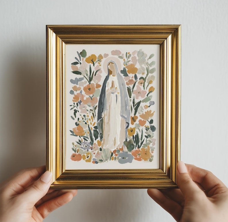 May include: A framed art print featuring a religious figure surrounded by colorful flowers. The artwork is in a gold frame and held by hands. The figure is wearing a white robe and a blue veil.