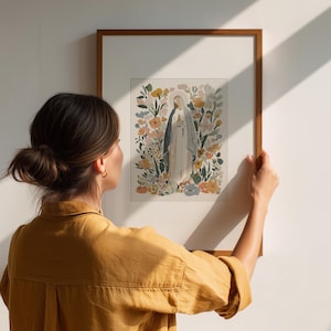 May include: A framed watercolor print of a religious figure surrounded by flowers. The artwork features soft colors and is displayed in a wooden frame. The image is being held up against a white wall.
