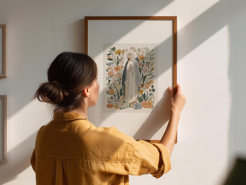 May include: A framed watercolor print of a religious figure surrounded by flowers. The artwork features soft colors and is displayed in a wooden frame. The image is being held up against a white wall.