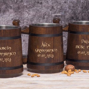 Puede incluir: Tres jarras de cerveza de madera con interiores de acero inoxidable. Cada jarra está grabada con un nombre, la palabra "Groomsmen" o "Best Man" y una fecha en letras doradas. Las jarras son de color marrón oscuro con bandas negras.
