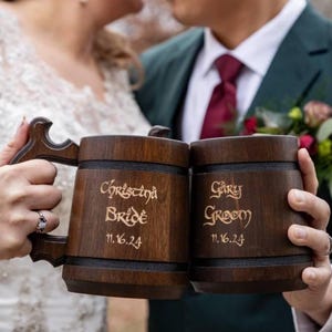 Könnte beinhalten: Zwei dunkelbraune Holzkrüge mit geschnitzten Griffen. Der linke Krug ist mit "Christina Bride 11.16.24" und der rechte Krug mit "Gary Groom 11.16.24" in einer stilisierten Schrift graviert. Beide Krüge haben horizontale schwarze Bänder.