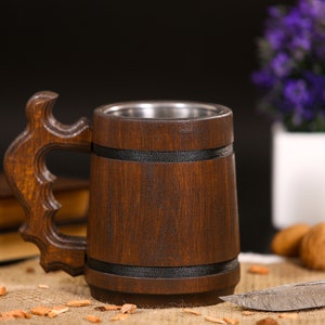 May include: A handcrafted wooden beer mug with a stainless steel interior. The mug features a dark brown finish, a decorative handle, and black bands. The mug is displayed on a burlap surface with a blurred background.