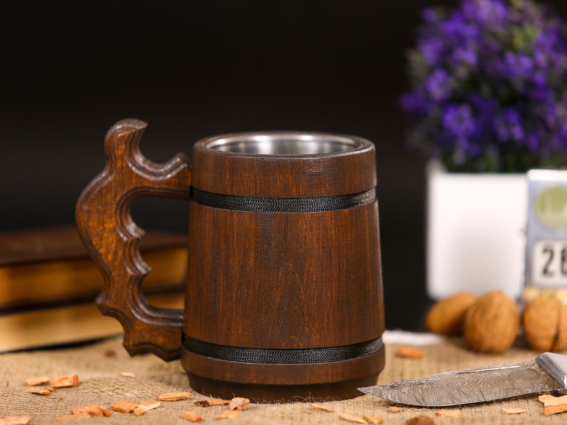 May include: A handcrafted wooden beer mug with a stainless steel interior. The mug features a dark brown finish, a decorative handle, and black bands. The mug is displayed on a burlap surface with a blurred background.
