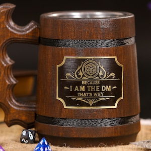 May include: Dark brown wooden beer mug with a decorative handle and a metal interior. The mug has a gold-coloured plaque with the text "BECAUSE I AM THE DM THAT'S WHY". Dice are scattered in front of the mug.