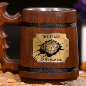 Könnte beinhalten: Ein dunkelbrauner Bierkrug aus Holz mit einem dekorativen Griff. Der Krug hat eine Metallauskleidung und eine goldene Plakette mit dem Text "THE TEARS OF MY PLAYERS" und einer Würfelgrafik.