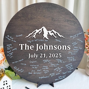 May include: A dark brown wooden wedding guest book with the names "The Johnsons" and the date "July 21, 2025" in white. A white mountain range is at the top. Guests have written messages around the edge.