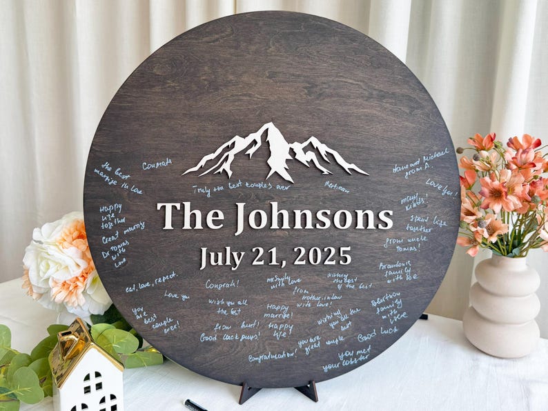 May include: A dark brown wooden wedding guest book with the names "The Johnsons" and the date "July 21, 2025" in white. A white mountain range is at the top. Guests have written messages around the edge.