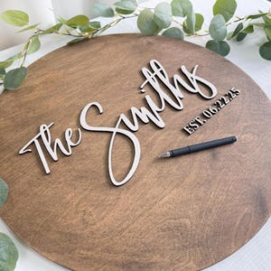 May include: A round wooden sign with the words "The Smiths" in a cursive font. The date "EST. 06.22.25" is also displayed. A black pen rests on the sign, which is surrounded by greenery.