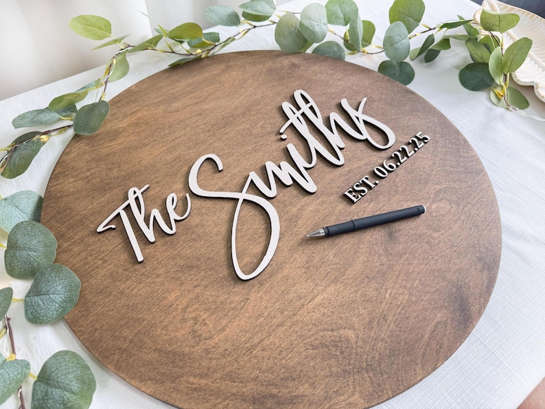 May include: A round wooden sign with the words "The Smiths" in a cursive font. The date "EST. 06.22.25" is also displayed. A black pen rests on the sign, which is surrounded by greenery.