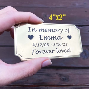 Puede incluir: Placa rectangular de latón, de aproximadamente 10 cm x 5 cm, con esquinas redondeadas. La placa dice "In memory of Emma", con un símbolo de corazón a cada lado del nombre, y las fechas "4/12/06 - 3/10/23". El texto "Forever loved" está debajo.