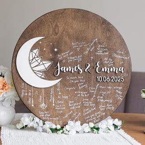 May include: A round wooden wedding guest book with a crescent moon and crystal design. The names "James & Emma" and the date "10.06.2025" are displayed. Guests have written well wishes around the edges.