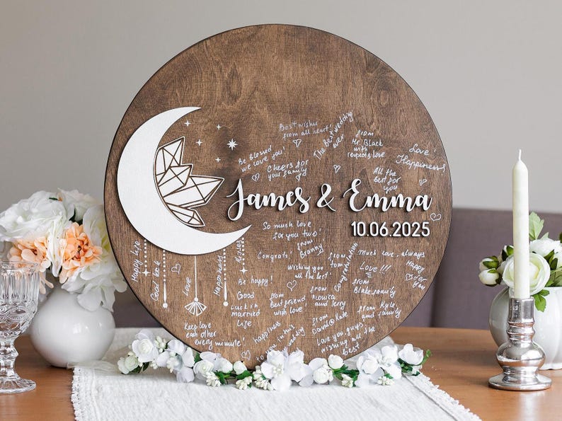 May include: A round wooden wedding guest book with a crescent moon and crystal design. The names "James & Emma" and the date "10.06.2025" are displayed. Guests have written well wishes around the edges.