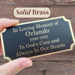 Op de afbeelding: Een rechthoekige zwarte messing plaquette met gouden rand, vastgehouden door een hand. De plaquette leest "In Loving Memory of Orlando 2006-2023 In God's Care and Always In Our Hearts." De tekst "Solid Brass" staat erboven.