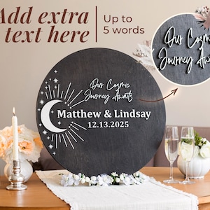May include: A round, dark gray wooden sign with white text that reads "Our Cosmic Journey Awaits" and the names "Matthew & Lindsay" with the date "12.13.2025". The sign features a crescent moon and star design.