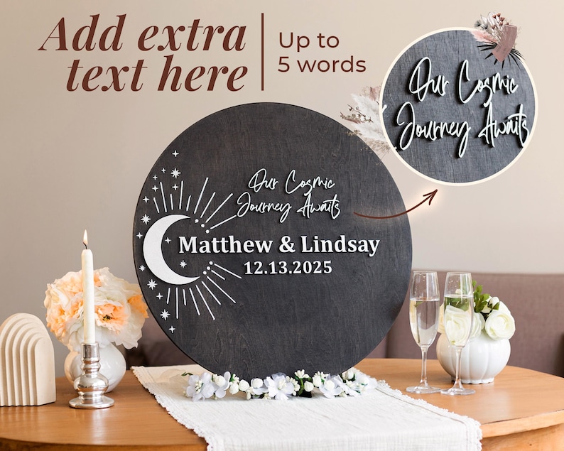 May include: A round, dark gray wooden sign with white text that reads "Our Cosmic Journey Awaits" and the names "Matthew & Lindsay" with the date "12.13.2025". The sign features a crescent moon and star design.