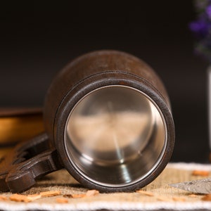 May include: A dark brown wooden beer mug with a stainless steel interior. The mug has a sturdy handle and is set against a dark background with a hint of a book and a decorative knife.