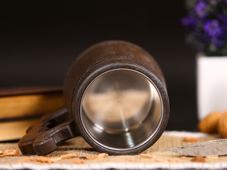 May include: A dark brown wooden beer mug with a stainless steel interior. The mug has a sturdy handle and is set against a dark background with a hint of a book and a decorative knife.