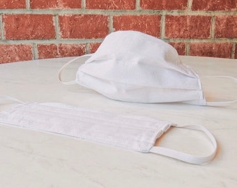 Puede incluir: Dos mascarillas de tela blancas con lazos elásticos para las orejas. Las mascarillas están dobladas y colocadas planas sobre una superficie de mármol blanco.