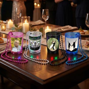 May include: Four illuminated shot glasses with attached beaded necklaces. The glasses are pink, clear, black, and blue, each displaying a different image: a woman, three dogs, a couple, and two silhouettes. The glasses are on a wooden table.