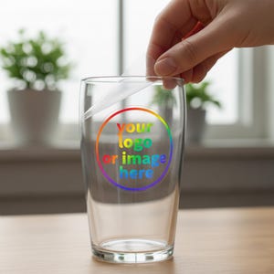 May include: A clear glass pint glass with a rainbow-colored circle design that reads "your logo or image here." A hand is peeling off a protective film from the glass. The glass is on a wooden surface.