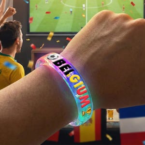 May include: A clear, glowing bracelet with the word "BELGIUM" in colorful letters. The bracelet features a soccer ball graphic and a trophy icon. The image shows the bracelet on a person's wrist, with a soccer game on TV in the background.