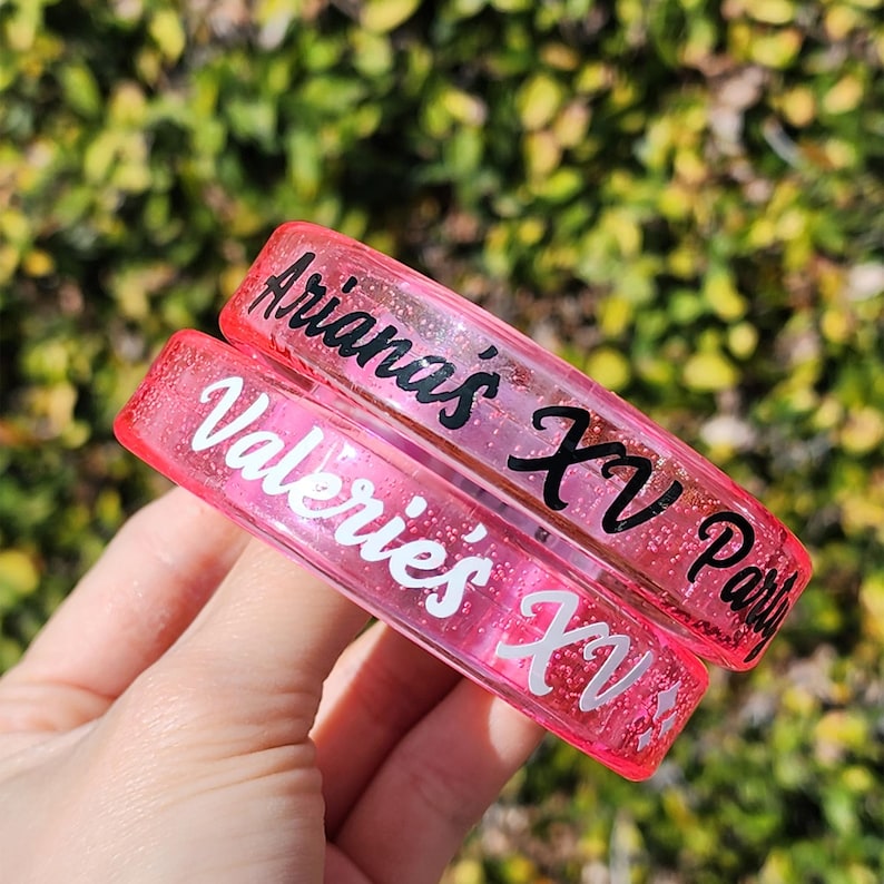 May include: Two pink, glitter-filled bracelets with text. One reads "Arianas XV Party" in black script, the other reads "Valerie's XV" in white. The bracelets are held against a blurred green background.