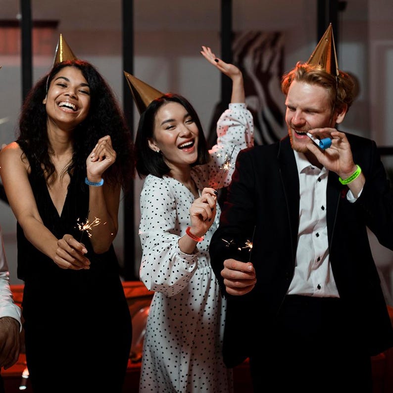 May include: A group of people celebrating, wearing party hats and holding sparklers. One person is wearing a black suit, and another is wearing a white dress with black polka dots. The people are smiling and laughing, with a festive atmosphere.