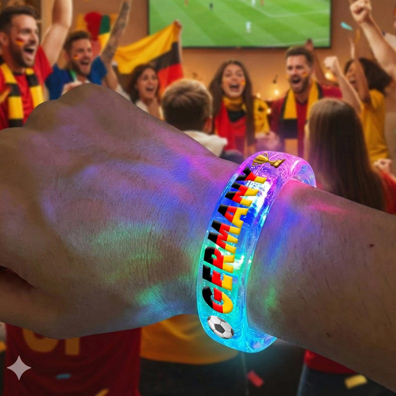 May include: A clear, glowing LED bracelet with the word "GERMANY" in black, yellow, and red, with a soccer ball graphic. The bracelet is worn on a wrist, with a crowd cheering in the background.
