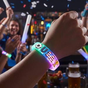 May include: A light-up wristband with the word "USA" and a soccer ball graphic. The wristband has a colorful design with the American flag and a trophy. The wristband is worn on a person's wrist.
