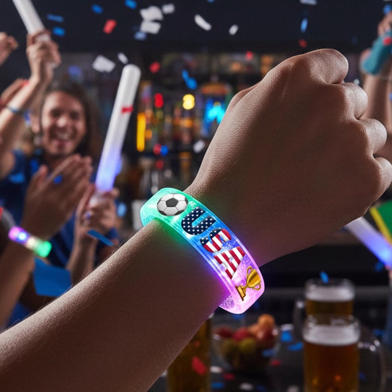 May include: A light-up wristband with the word "USA" and a soccer ball graphic. The wristband has a colorful design with the American flag and a trophy. The wristband is worn on a person's wrist.