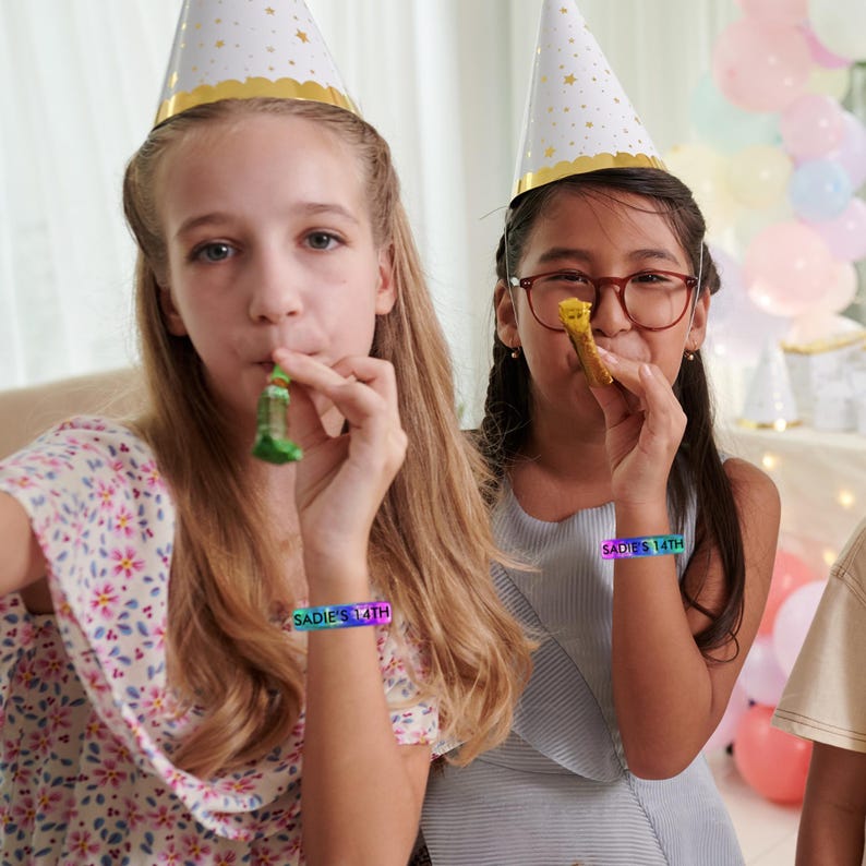 May include: Two young people wearing party hats and bracelets that read "SADIE'S 14TH" are blowing party favors. One person is wearing a floral print top, and the other is wearing glasses and a light blue top. Balloons are visible in the background.