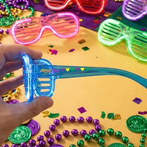 May include: A close-up of a pair of blue light-up sunglasses with the text "Mardi Gras" and heart designs. Other pairs of light-up sunglasses in pink, red, and green are visible, along with Mardi Gras beads and confetti.