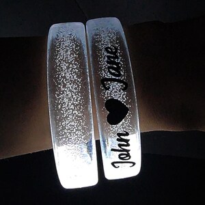 May include: Two clear, glowing bracelets with the names "John" and "Jane" in black script, along with a black heart. The bracelets are illuminated with tiny white specks, creating a sparkling effect. The background is dark.