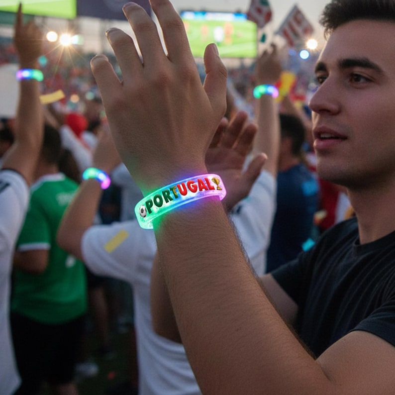 May include: A person wearing a glowing LED bracelet with the word "PORTUGAL" in red letters. The bracelet has a colorful, illuminated border. Other people in the background are also wearing similar bracelets. The scene appears to be at a sporting event.