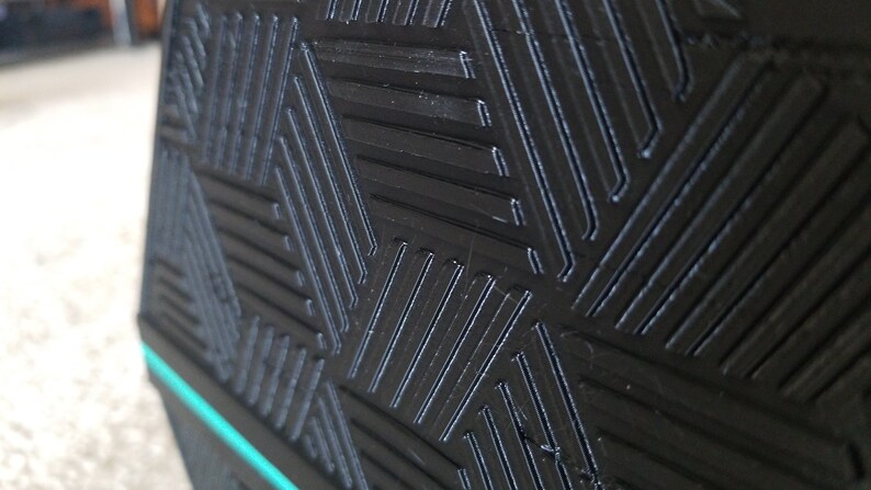 May include: Close-up of a black object with a geometric pattern of diagonal lines. The lines are raised, creating a textured surface. A thin, bright teal stripe accents the edge of the object. The overall design is modern and minimalist.