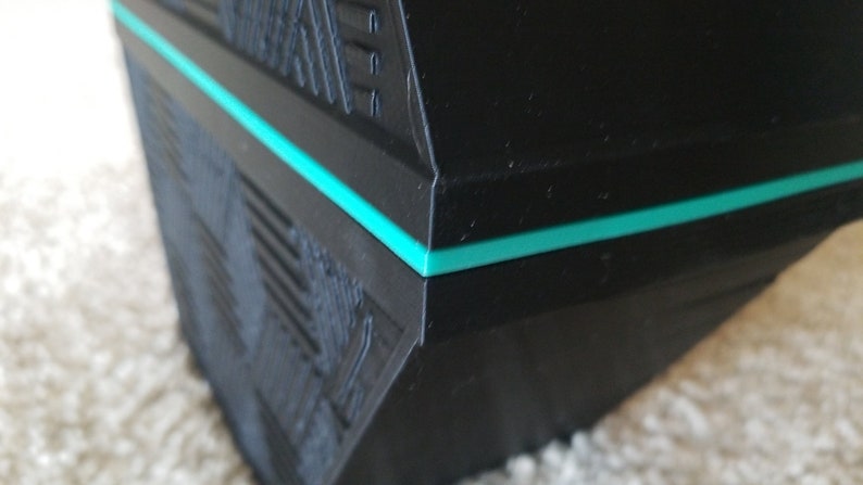 May include: Close-up of a black, geometric object with a textured surface and a thin, teal-colored stripe. The object has a modern, angular design, with visible ridges and lines.