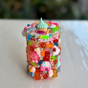 May include: A small, cylindrical container decorated with a variety of colourful, miniature sweets, beads, and decorative elements. The container has a light green swirl on top and is adorned with fruit and dessert-themed embellishments.