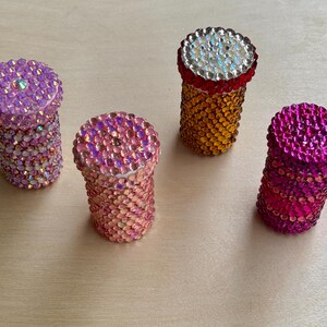 Op de afbeelding: Vier kleine, ronde containers met deksels bedekt met kleurrijke strass steentjes. De containers zijn roze, oranje en twee tinten paars.