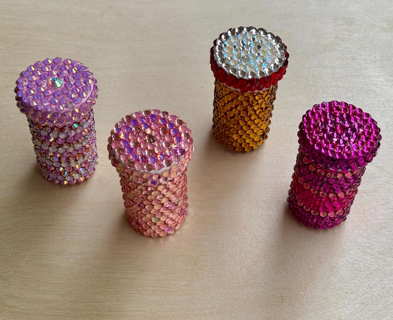 Op de afbeelding: Vier kleine, ronde containers met deksels bedekt met kleurrijke strass steentjes. De containers zijn roze, oranje en twee tinten paars.