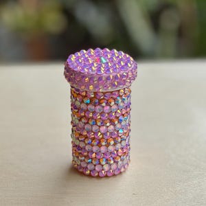 Op de afbeelding: Een kleine roze container bedekt met iriserende strass steentjes. De container is cilindrisch en heeft een deksel.