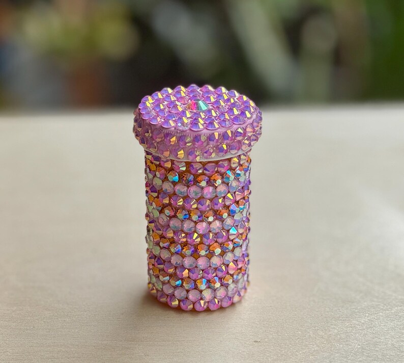 Op de afbeelding: Een kleine roze container bedekt met iriserende strass steentjes. De container is cilindrisch en heeft een deksel.