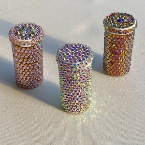 Könnte beinhalten: Drei zylindrische Behälter, die mit schillernden Strasssteinen bedeckt sind. Jeder Behälter hat einen weißen Deckel und steht auf einer weißen Oberfläche. Die Strasssteine reflektieren mehrere Farben und erzeugen einen funkelnden Effekt. Die Behälter sind unterschiedlich hoch.