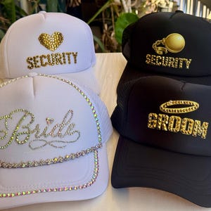 May include: Four trucker hats with mesh backs. Two white hats have the words "Security" and "Bride" in gold rhinestones. The other two black hats have the words "Security" and "Groom" in gold rhinestones.