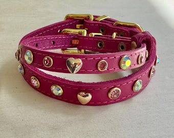 Puede incluir: Dos collares de perro de cuero fucsia apilados con hebillas doradas. Cada collar está adornado con pedrería iridiscente, pequeñas gemas rosas y detalles dorados en forma de corazón. Los collares están diseñados para mascotas.