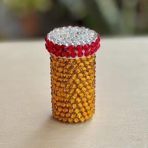 Op de afbeelding: Een kleine, ronde, gouden container met een rood-witte bovenkant bedekt met strass. De container is ongeveer 5 cm hoog en 2,5 cm in diameter.