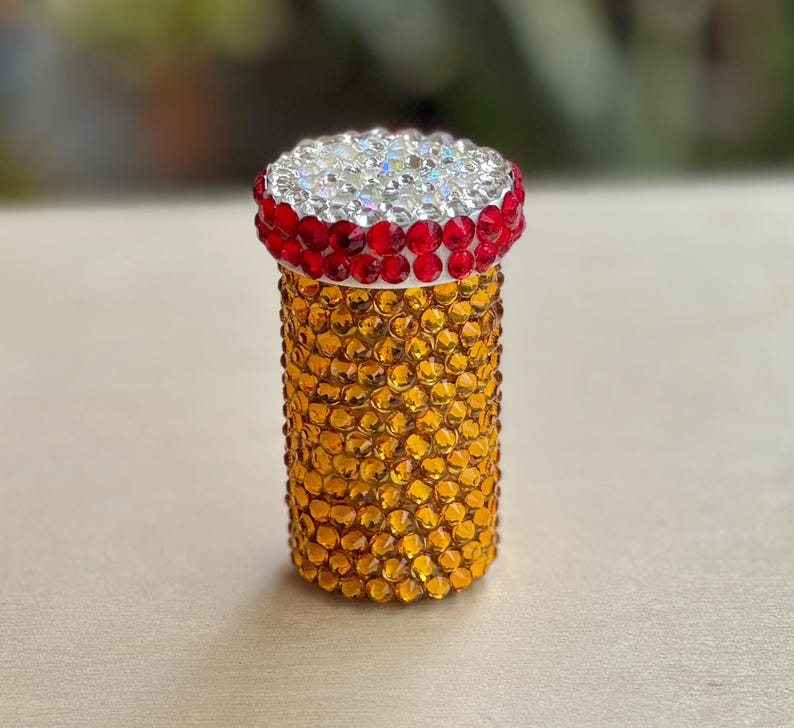 Op de afbeelding: Een kleine, ronde, gouden container met een rood-witte bovenkant bedekt met strass. De container is ongeveer 5 cm hoog en 2,5 cm in diameter.