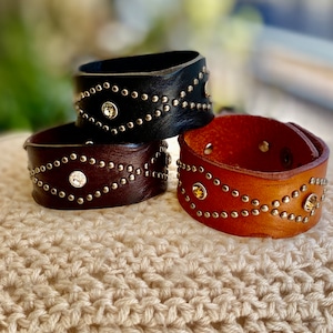 Op de afbeelding: Drie leren armbanden met zilveren studs en strass. De armbanden zijn op elkaar gestapeld. De bovenste armband is zwart, de middelste armband is bruin en de onderste armband is beige.