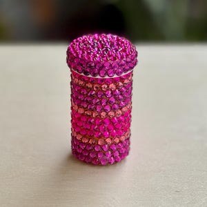 Op de afbeelding: Een roze en paarse juwelencontainer met een rond deksel. De container is bedekt met kleine, glinsterende strasssteentjes.